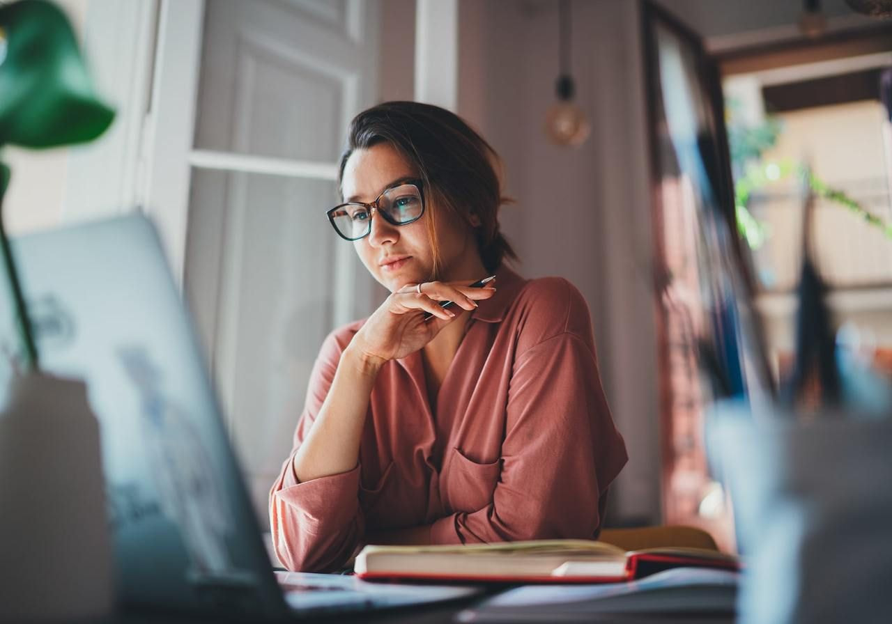 A woman wearing glasses is seated at a desk, working on a laptop.